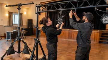 Two technicians clamping an LED PAR light to a truss goalpost on stands with safety cables, while a T-bar light stand has sandbags on its legs